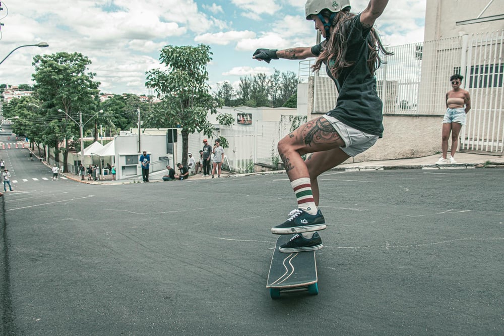 A young woman is skateboarding down a steep, urban street. She is wearing a helmet, black t-shirt, white shorts, and knee-high striped socks. Her arms are outstretched for balance as she maneuvers the skateboard, showcasing her tattoos on her legs. The background features several onlookers, a mix of people standing and sitting by the roadside.